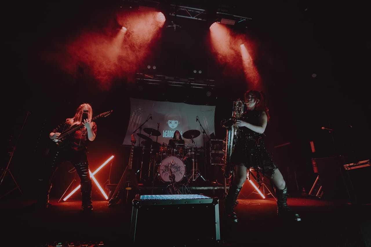 DEADGRACE live at The Ritz in San Jose: Grace Kelly plays baritone saxophone stage right, masked guitarist Sam Fairless plays stage left, and drummer Kyle Brian sits behind the kit in front of a DEADGRACE backdrop under red lights