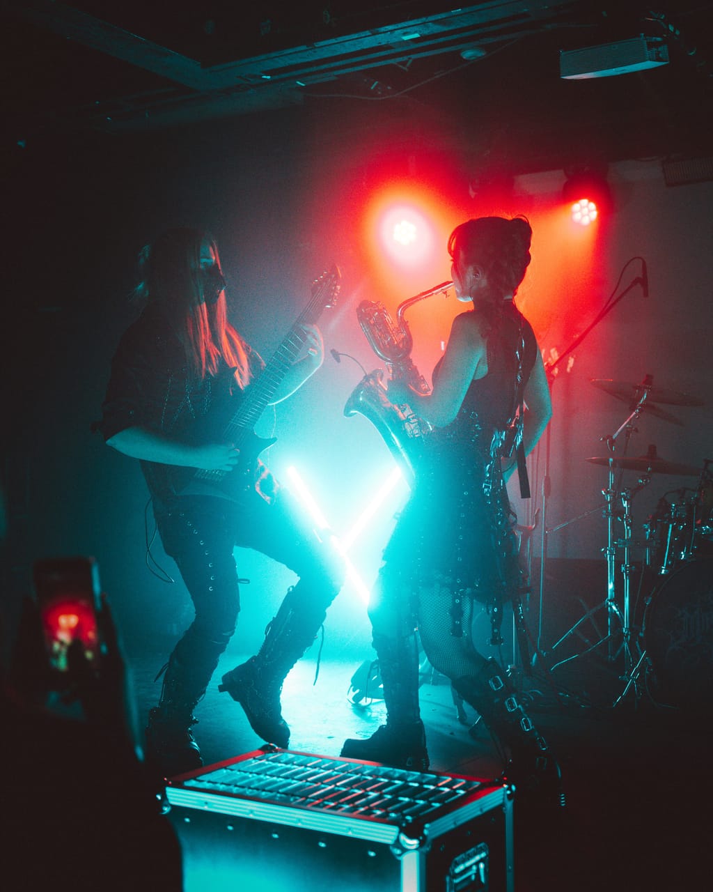 Guitarist Sam Fairless, long hair and a black mask, and Grace Kelly on saxophone play face to face in teal and red light at El Corazon, a road case at the front of the stage