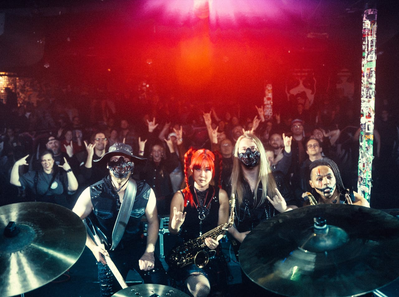 DEADGRACE pose on stage at El Corazon, photographed from behind the drum cymbals: Hampus Isaksson, Grace Kelly with her saxophone, Sam Fairless and Dane White, with the Seattle crowd throwing horns behind them and a sticker-covered pillar at right