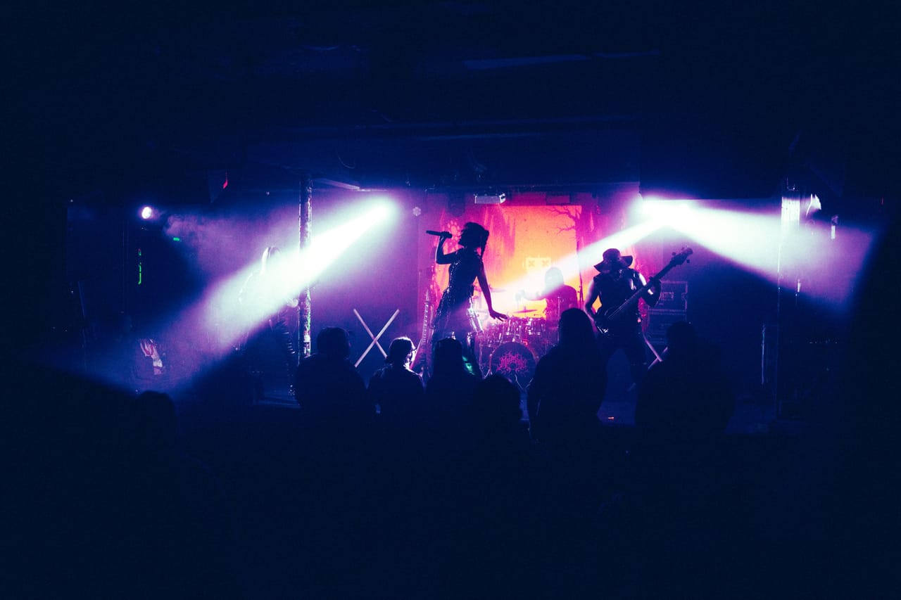 DEADGRACE on stage at El Corazon in Seattle seen from the back of the crowd: Grace Kelly silhouetted singing into a microphone and Hampus Isaksson in a hat on bass, white light beams cutting through haze over the audience