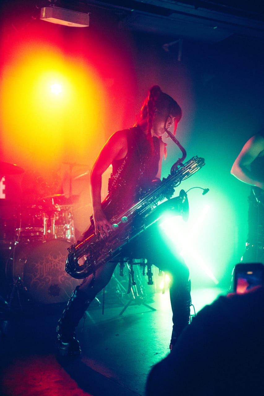 Grace Kelly, red braids tied up, leans into a large low saxophone under orange and green stage light at El Corazon, Seattle, the drum kit behind her and a phone raised in the crowd