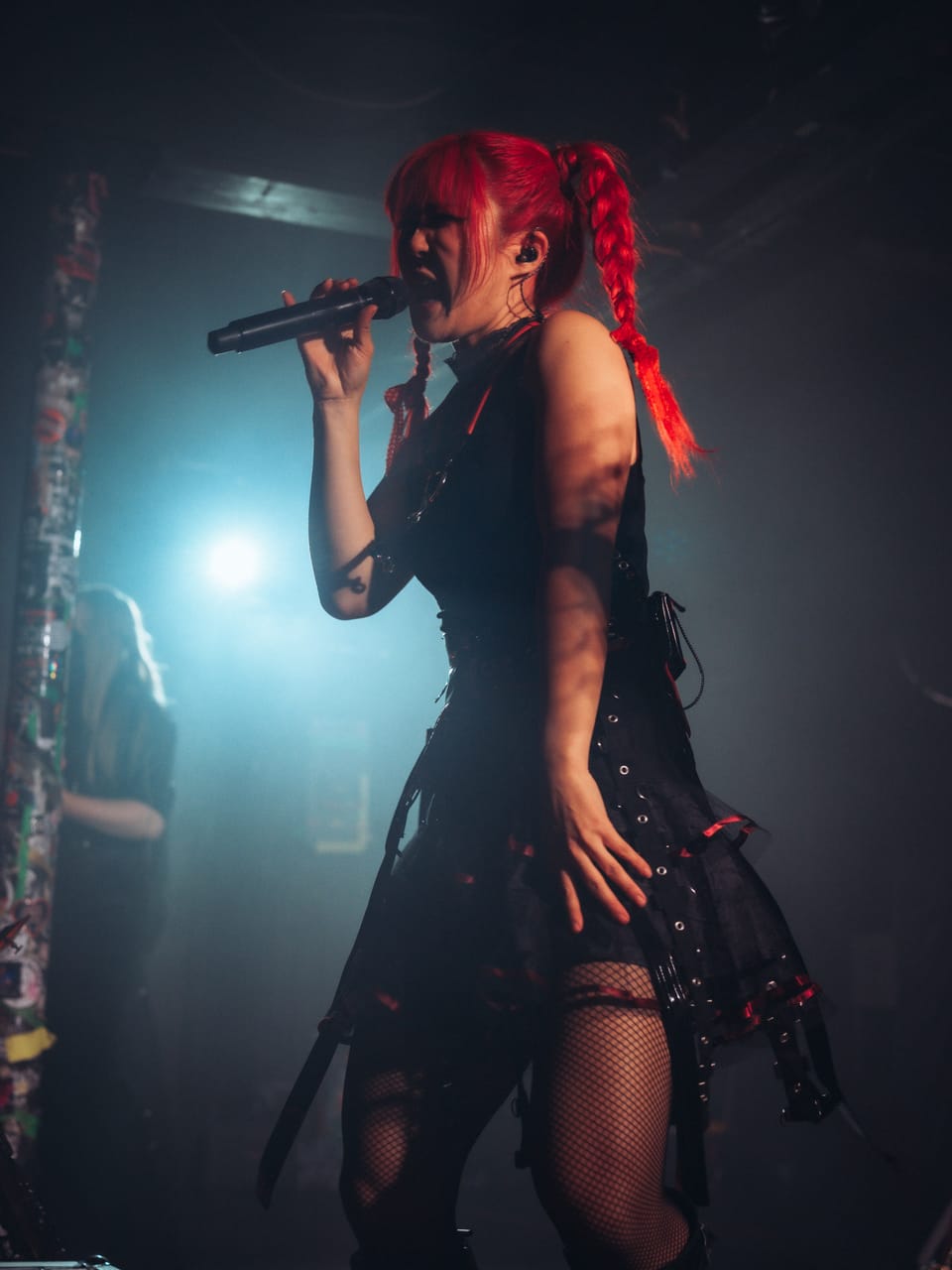 Grace Kelly sings into a handheld microphone through teal stage light and haze at El Corazon, red braids over her shoulder, Sam Fairless in the background