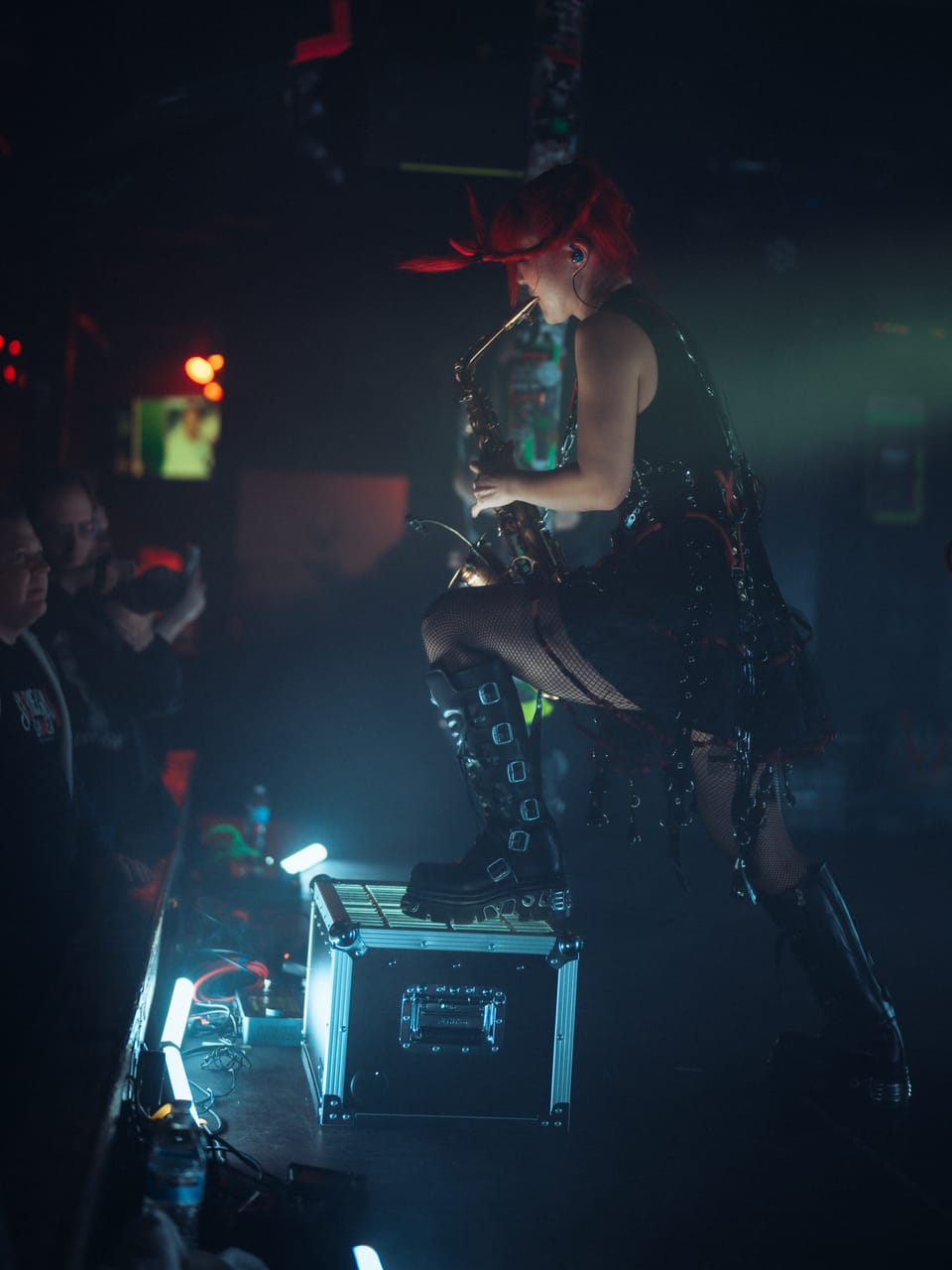 Grace Kelly, one boot on a road case at the edge of the stage, plays saxophone over the front row at El Corazon, seen from the side