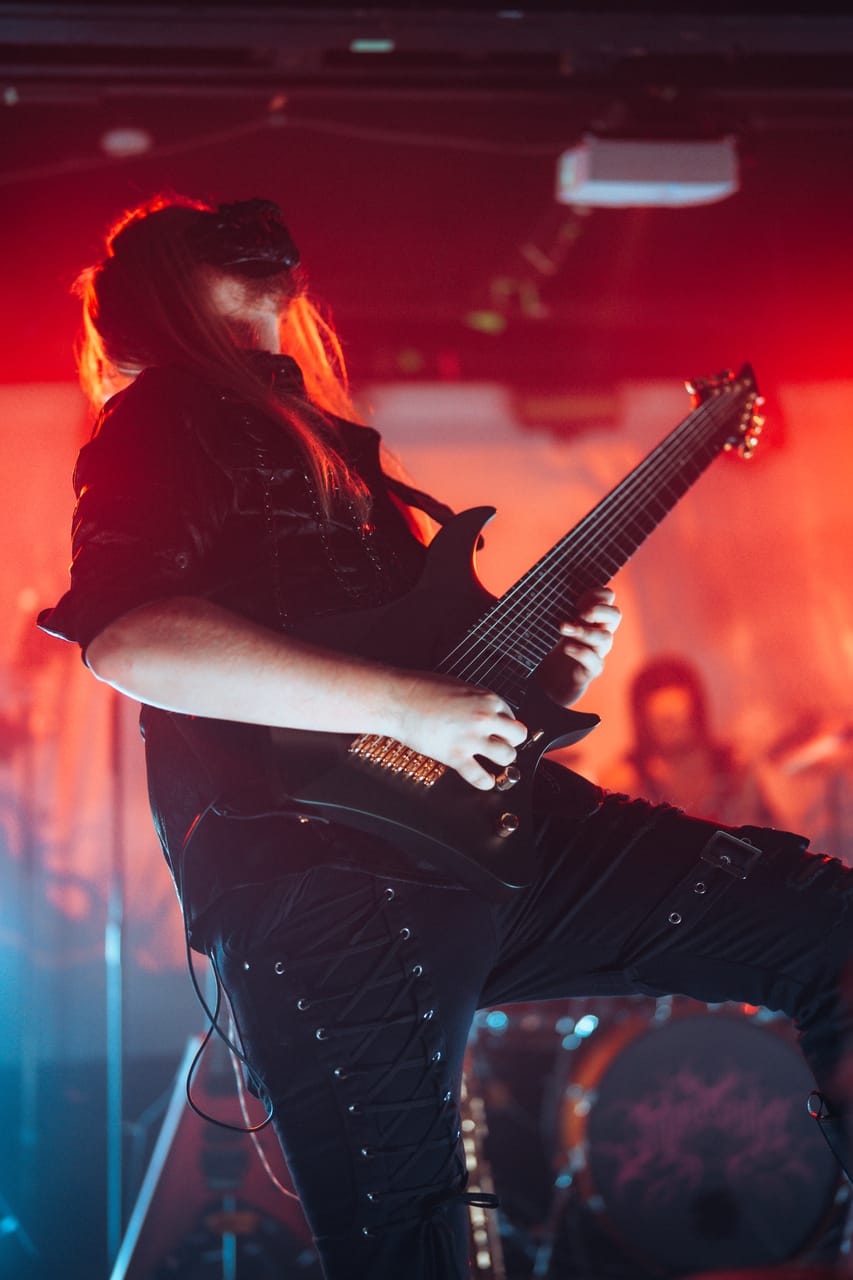 DEADGRACE guitarist Sam Fairless, long hair and a black mask, leans back playing an eight-string guitar under red light at El Corazon, Dane White on drums behind