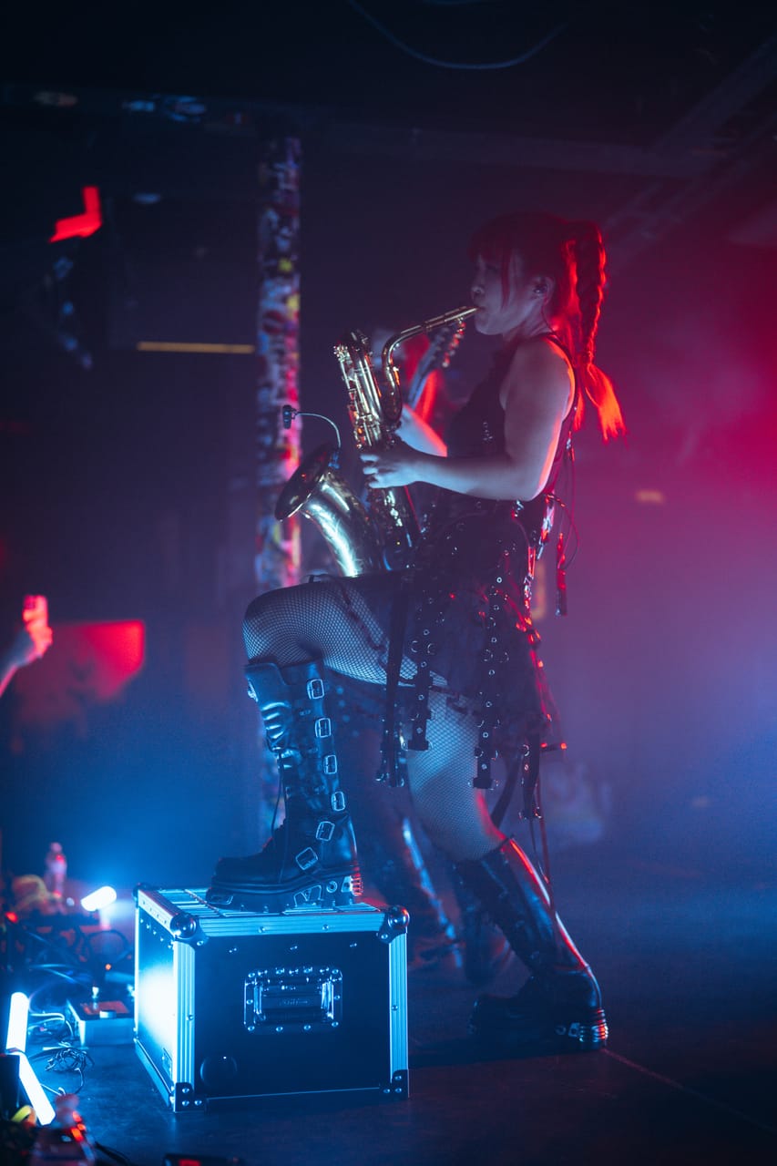 Grace Kelly plays saxophone with one boot on a road case under pink and blue light at El Corazon, Seattle
