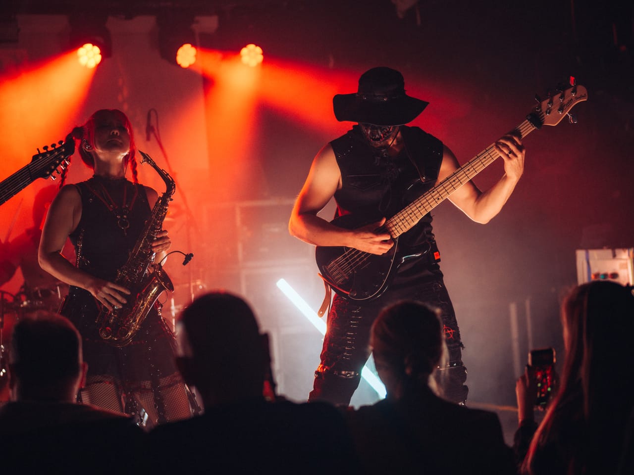 Grace Kelly plays saxophone beside bassist Hampus Isaksson, in a black hat, under red and orange lights at El Corazon, with fans watching and filming from the floor