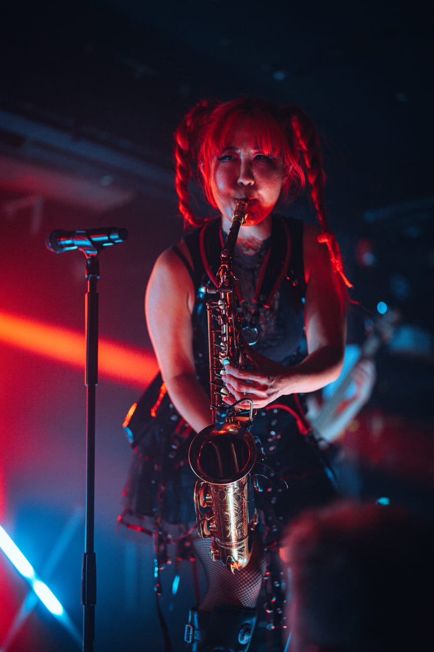 Grace Kelly plays saxophone straight at the camera under red light beside a microphone stand at El Corazon, Seattle