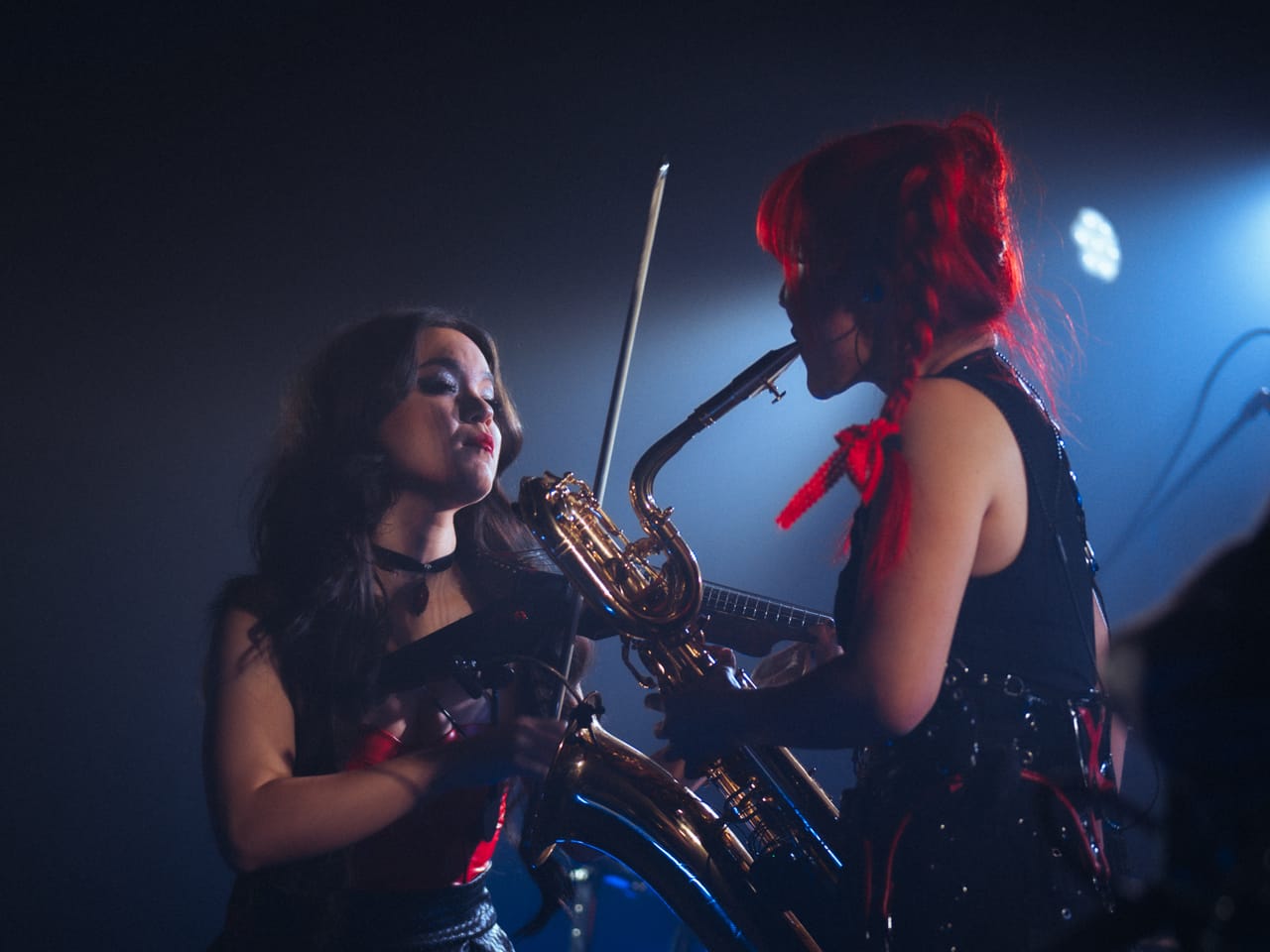 Mia Asano on electric violin and Grace Kelly on baritone saxophone play face to face under blue light on stage at El Corazon in Seattle