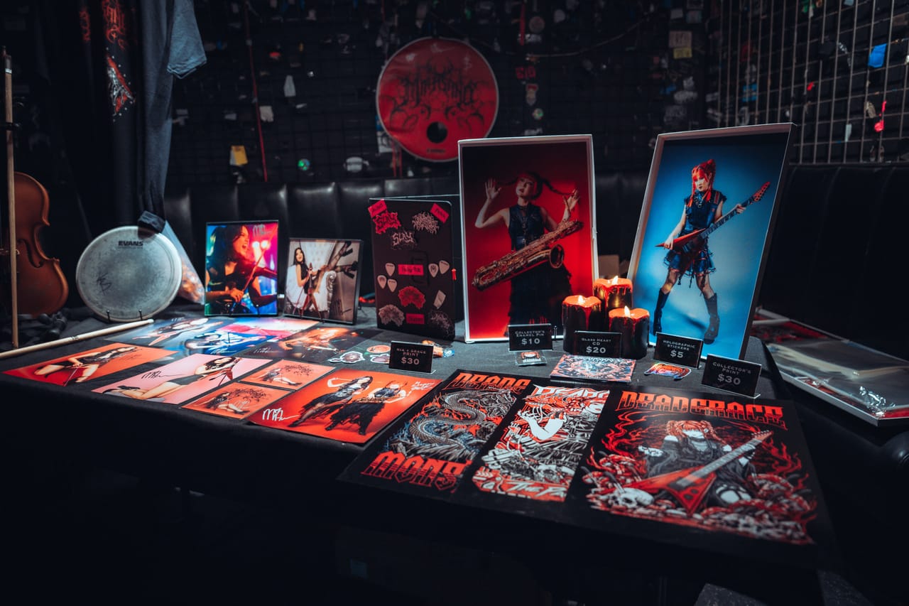The DEADGRACE merch table set up at El Corazon in Seattle: candles, posters, price cards, and framed prints of Grace Kelly with a baritone saxophone and a red flying-V guitar, a violin and a drumhead behind