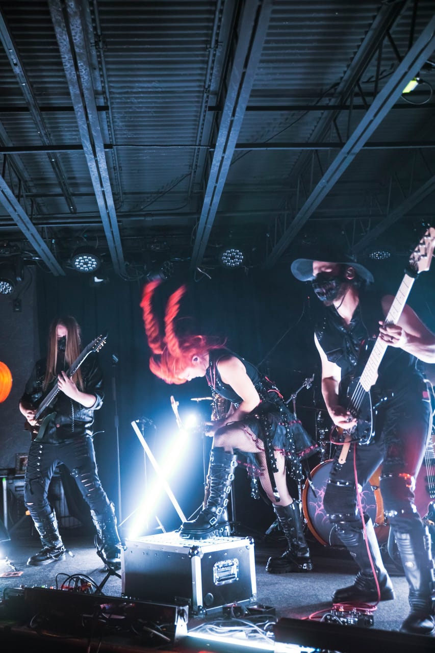 Grace Kelly headbangs with her red braids flying, one boot on a road case, between guitarist Sam Fairless and bassist Hampus Isaksson on stage at The District, Spokane