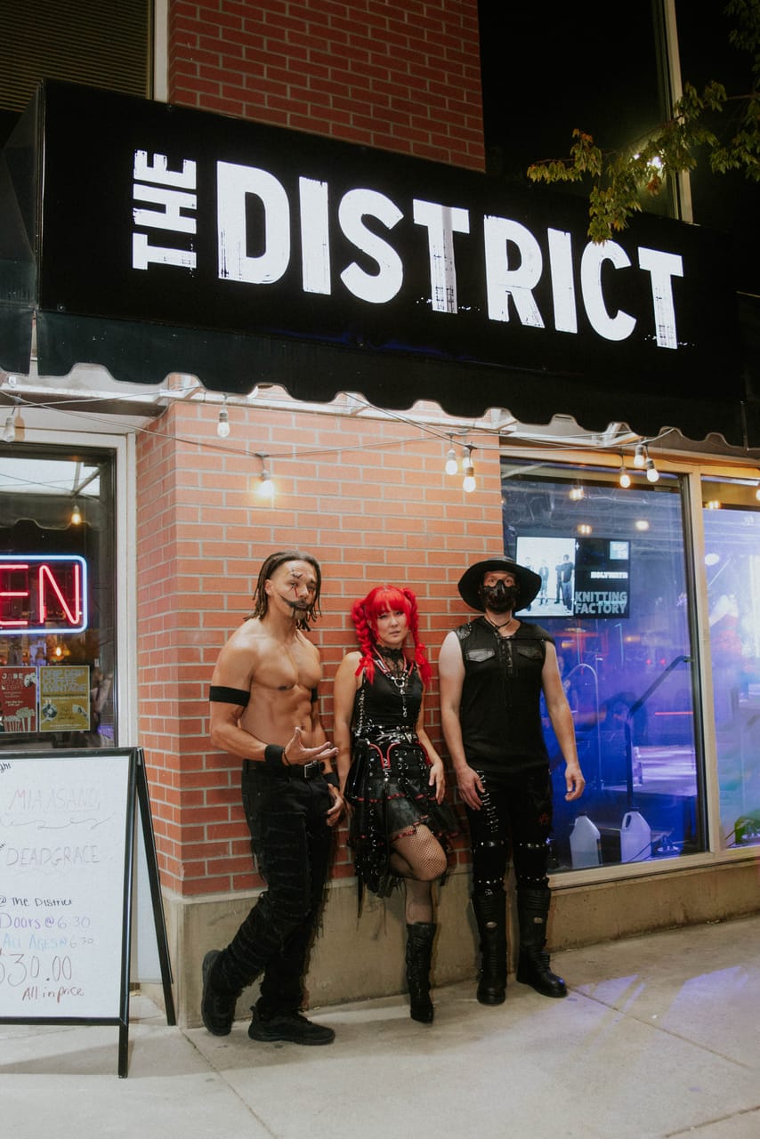 Dane White, Grace Kelly and Hampus Isaksson of DEADGRACE stand outside The District in Spokane under its sign, a chalkboard by the door listing Mia Asano and DEADGRACE, doors at 6:30, all ages