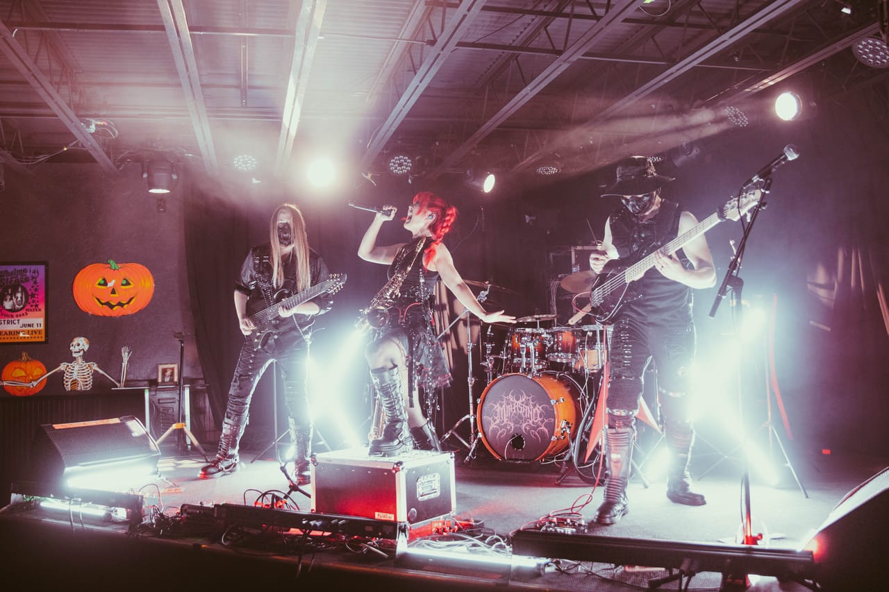 DEADGRACE on stage at The District in Spokane: Sam Fairless on guitar, Grace Kelly singing with a saxophone on its strap, and Hampus Isaksson on bass, a jack-o'-lantern and a skeleton at the side of the stage