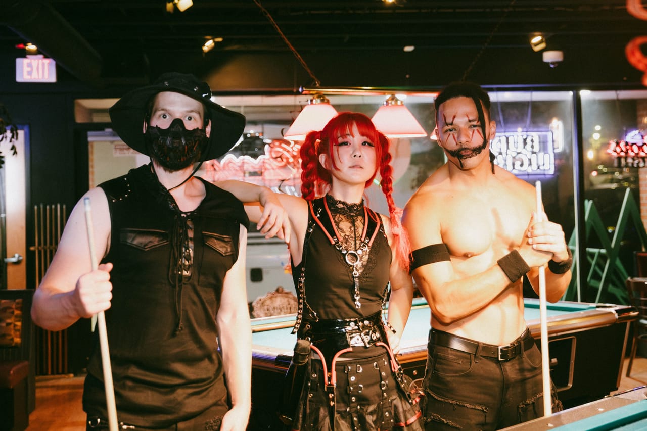 Hampus Isaksson in a hat and mask, Grace Kelly, and Dane White in face paint hold pool cues beside a pool table at The District in Spokane after the show