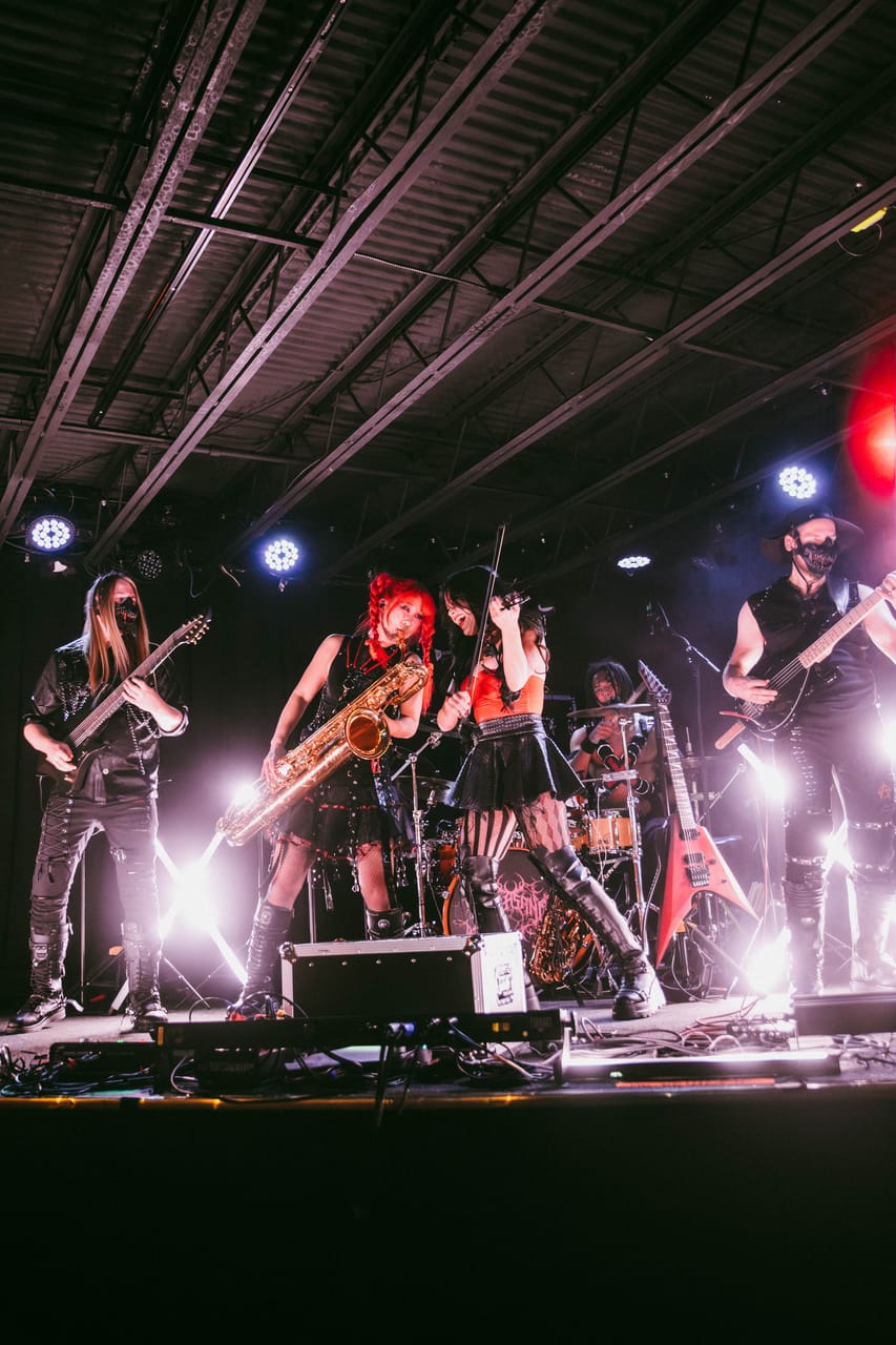 Grace Kelly on saxophone and Mia Asano on violin play side by side between Sam Fairless and Hampus Isaksson on stage at The District in Spokane, a red flying-V guitar on its stand