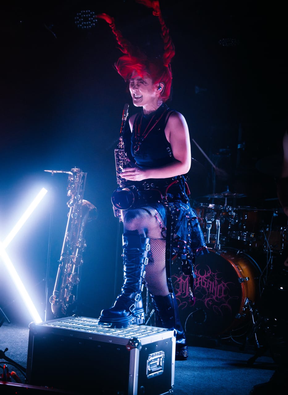 Grace Kelly plays saxophone standing on a road case, a braid flying above her head, in blue light at The District, Spokane, a baritone saxophone on its stand behind her
