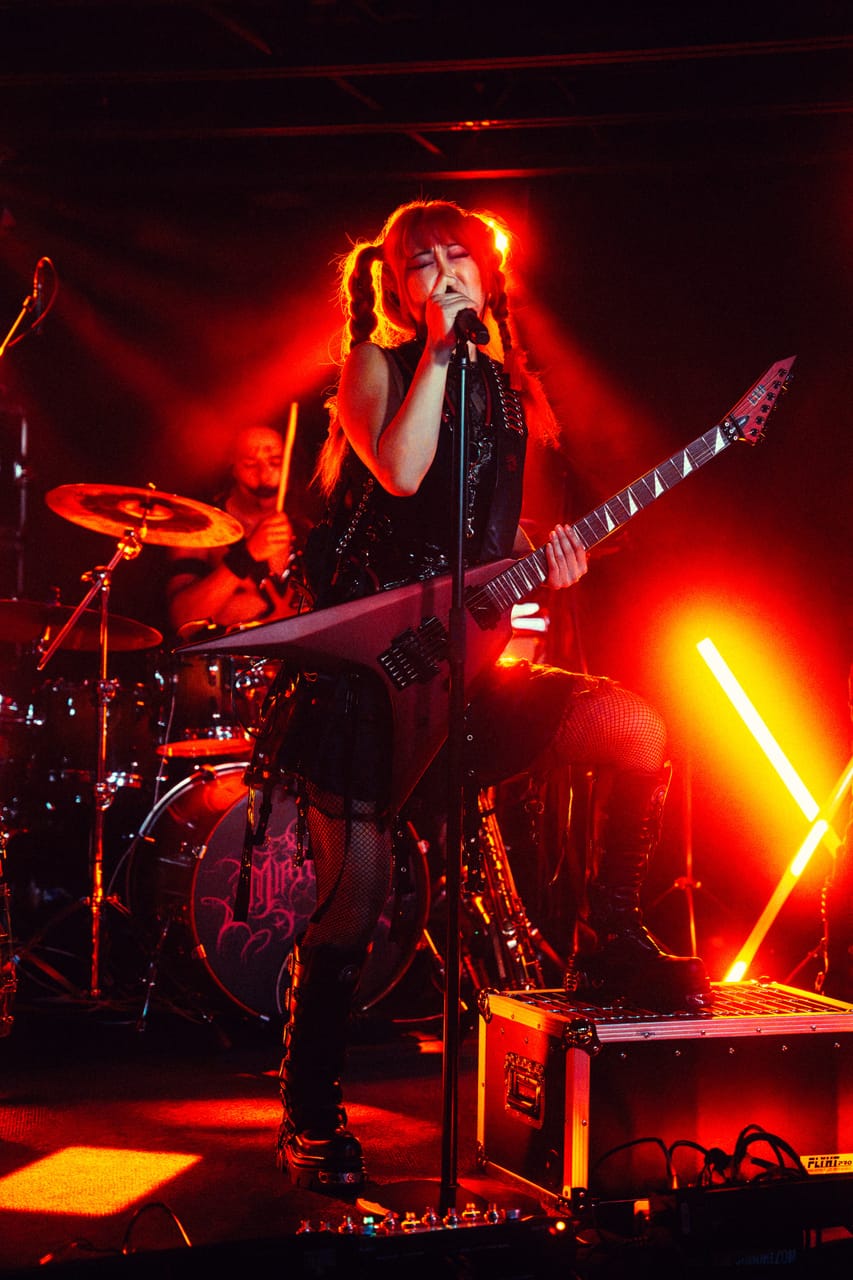 Grace Kelly sings with a red flying-V guitar, one boot on a road case, under red light beside a glowing neon X at The District, Spokane