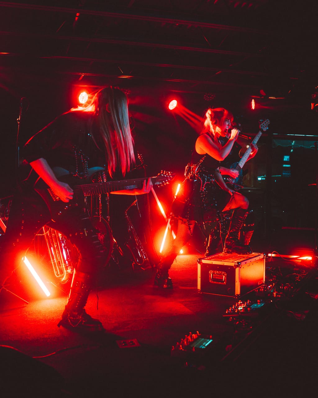 Guitarist Sam Fairless, long hair over his face, plays in the foreground as Grace Kelly sings with a knee up on a road case, the stage washed in red at The District in Spokane