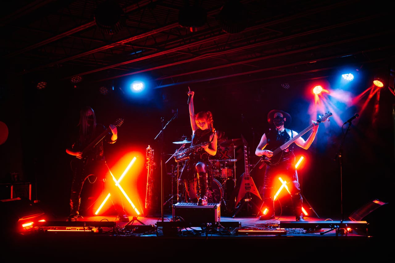 Wide shot of DEADGRACE on stage at The District, Spokane: Sam Fairless on guitar, Grace Kelly with a fist in the air and a saxophone on its strap, and Hampus Isaksson in a hat on bass, neon X lights glowing red