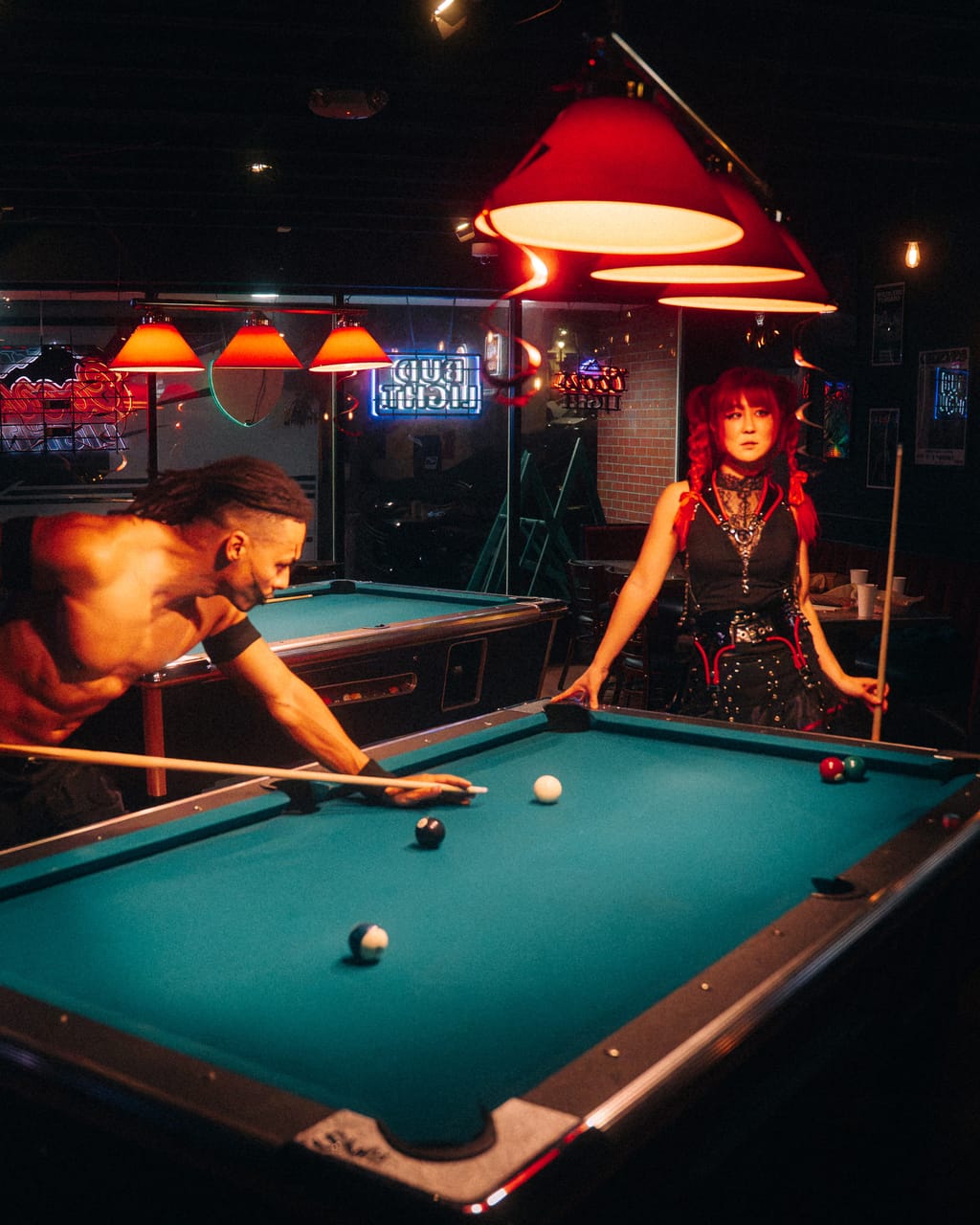 Dane White leans over a pool table lining up a shot while Grace Kelly waits with her cue under red lamps at The District in Spokane